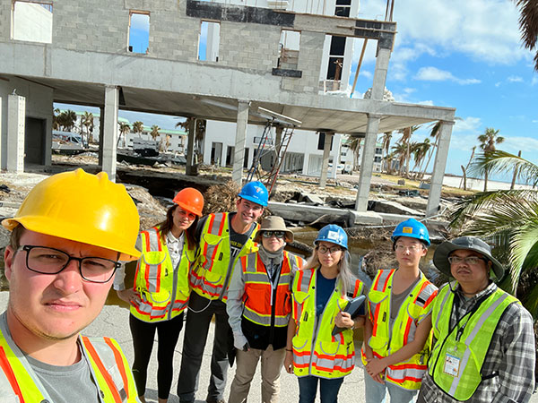 a group of researchers in hard hats standing in front a hurricane-damaged building