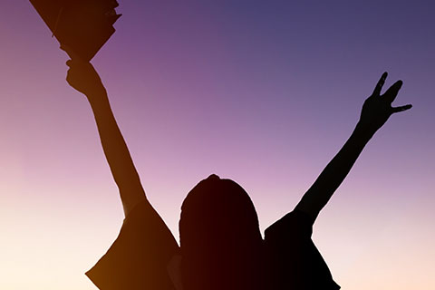 A silhouette of a student holding up a graduation cap 