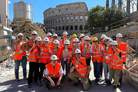 A group of students wearing orange safety vests posing in front of the Roman Colosseum 