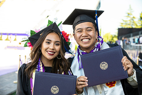 Two students in graduation gowns holding diplomas in their hands