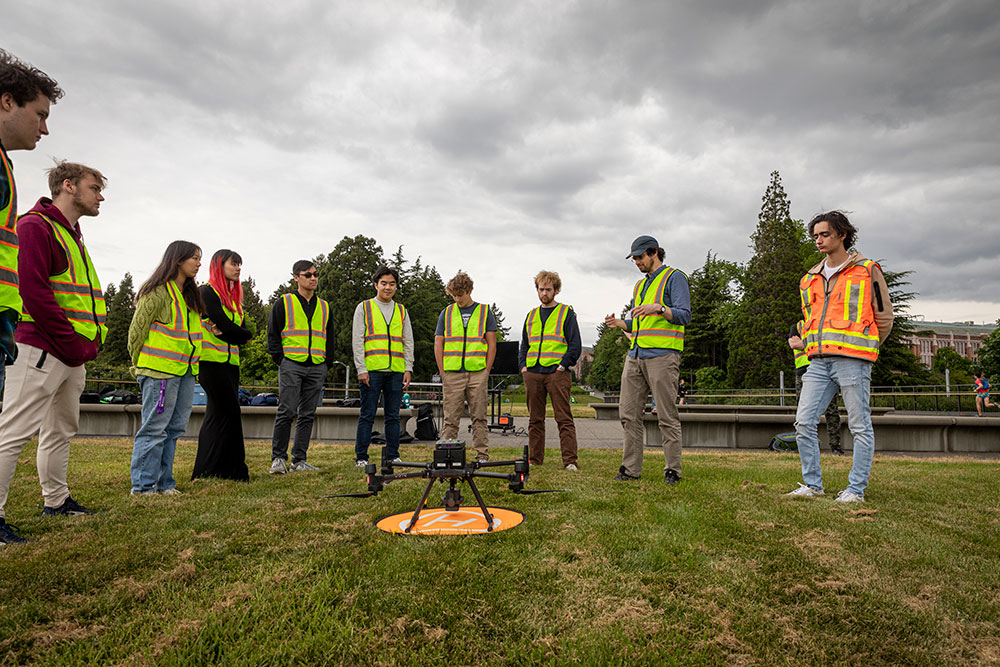 A group of students wearing safety vests standing around a drone on a lawn