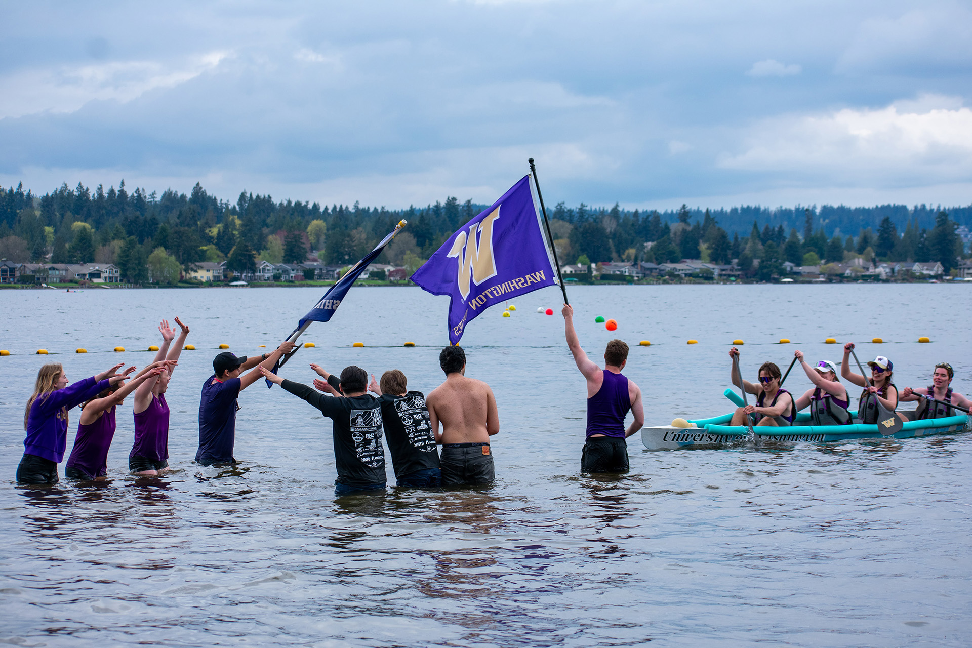 A line of students wade waist-deep into a lake, raising their arms and waving flags as a canoe team paddles toward them