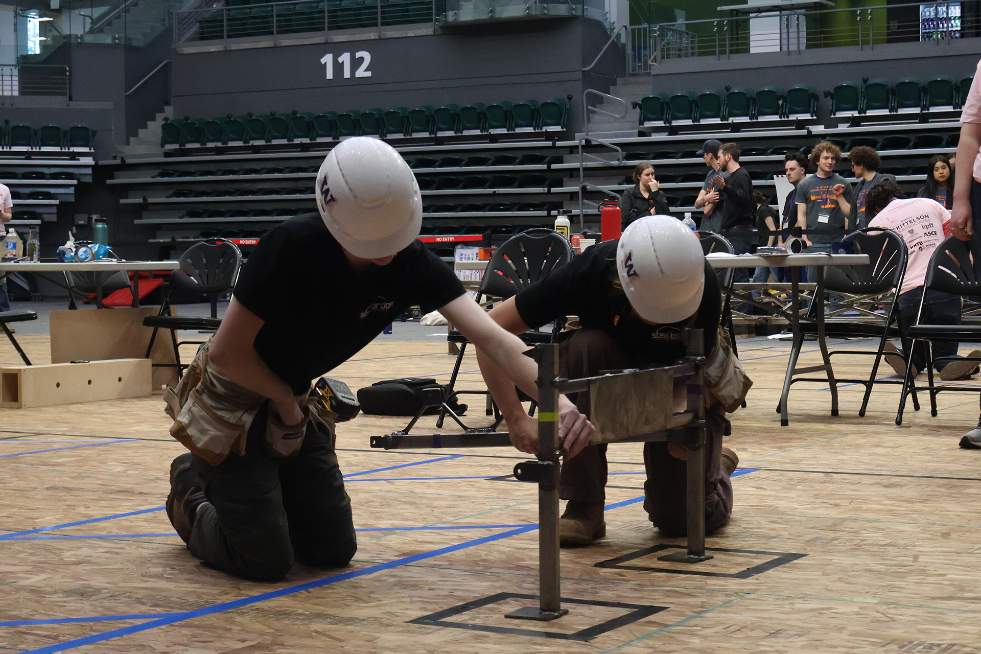 Two students in white hard hats assembling steel bridge components with tool belts at their sides.