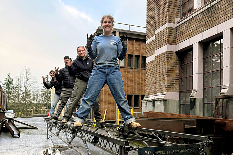 Four students stand on top of a fully assembled steel truss bridge outside a brick building, smiling and gesturing at the camera
