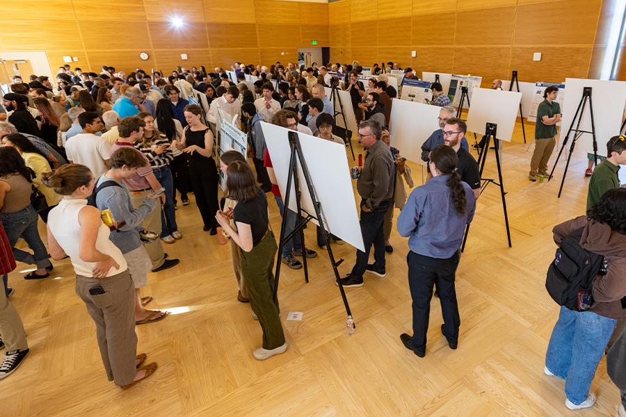 A large crowd mingles among rows of poster easels in a wood-paneled event hall