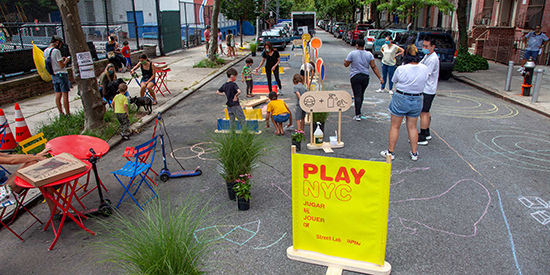 Street in New York City closed to cars and turned into a play street with kids, chalk drawings and “PLAY NYC” sign.