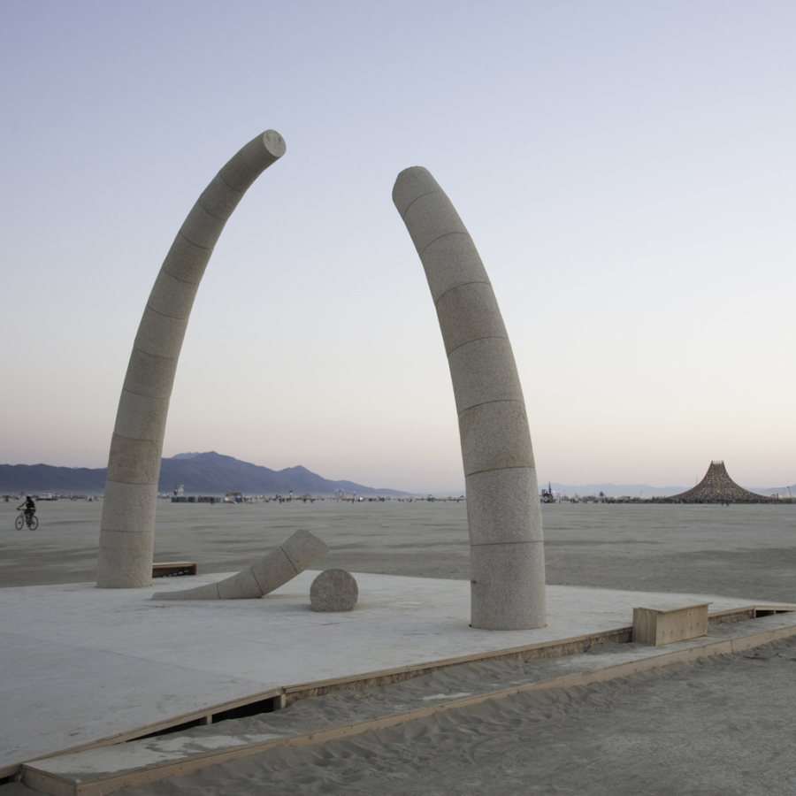 Two tall granite columns curve toward each other but do not meet, with a missing segment and a cylindrical stone piece resting on the ground between them, set against a desert landscape at dusk.