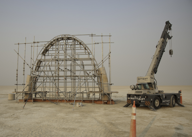 A partially assembled stone arch surrounded by scaffolding on a flat desert landscape, with a crane parked alongside.