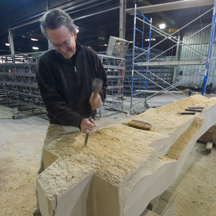 Richard Rhodes smiling as he carves a large piece of stone with a hammer and chisel in a workshop.