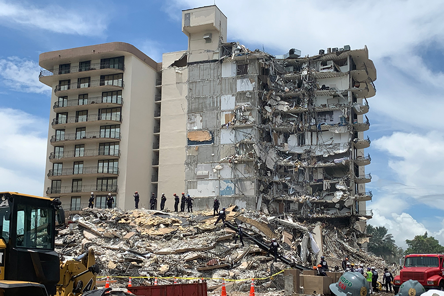 A large condominium that has partially collapsed, and people with helmets amidst the rubble.