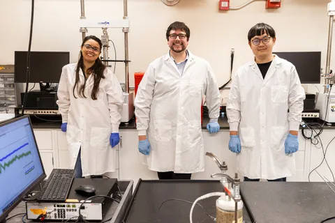 Three people in lab coats and gloves standing in a laboratory with equipment behind them.