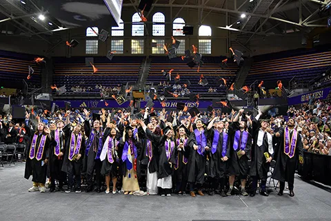 A large group of CEE graduates in black gowns and purple stoles toss their caps into the air inside Alaska Airlines Arena.