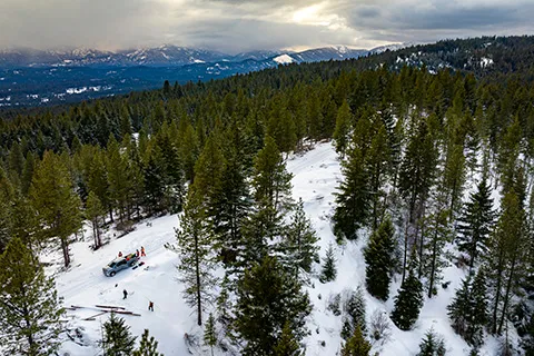 An aerial photo of a snowy forest with a mountain range in the background. In the foreground, several small figures stand next to a pickup truck.