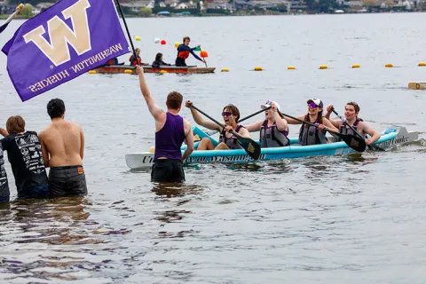 UW Concrete Canoe paddlers in Grand Coulee approach shore as a teammate waves a UW flag in shallow water.