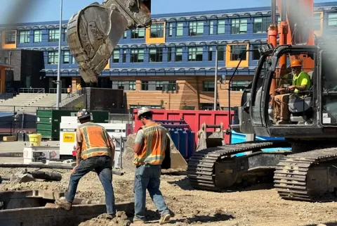 Two construction workers walk in front of a bulldozer on a construction site.