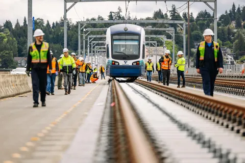 Construction workers walk on either side of a lightrail train on a track.